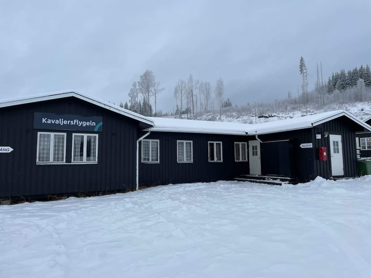Stuga på området med mörkgrå träfasad, flera fönster och entré under tak