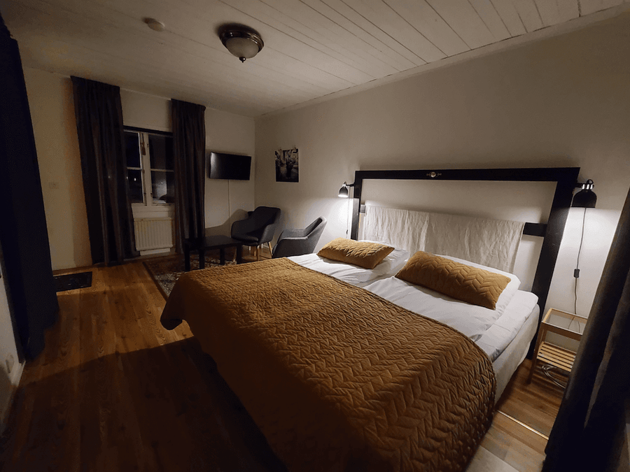 Sovrum med dubbelsäng, beige täcke och två kuddar, trägolv och gardiner